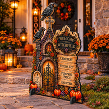 Load image into Gallery viewer, A decorative Halloween yard sign shaped like a witchfield tombstone with two crows perched on top, displayed in front of a warmly lit autumnal house. The sign features faux-stone textures, carved details, faux pumpkins, and faux flowers, with text reading 'WELCOME TO WITCHFIELD' and additional seasonal phrases.