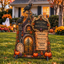 Load image into Gallery viewer, A decorative Halloween yard sign shaped like a gothic graveyard house with two crows, tombstones, pumpkins, autumn leaves, and a sign reading “Welcome to Witchfield” and “Cemetery Witch’s Cot Pumpkin Patch Haunted,” displayed on a grassy lawn in front of a house.