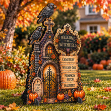 Load image into Gallery viewer, A decorative Halloween yard sign shaped like a haunted witch house with crows, autumn flowers, pumpkins, and a sign reading 'Welcome to Witchfield' and additional seasonal text, displayed on grass in an outdoor yard with fall foliage.