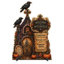 Load image into Gallery viewer, Halloween decorative witchfield cemetery sign featuring a gothic tombstone structure with two black crows, a central iron gate, orange pumpkins, flowers, and a wooden-style sign reading 'Welcome to Witchfield' with additional cemetery-themed phrases.