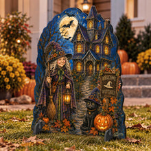 Load image into Gallery viewer, A Halloween yard sign shaped like a tombstone stands on grass in front of a house. The design shows a witch in a purple robe holding a broom beside a haunted house under a full moon, with a black cat, pumpkins, autumn leaves, and a sign reading “Welcome Witches.