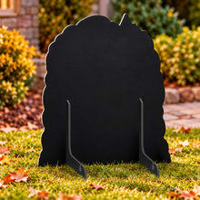 Load image into Gallery viewer, A black cardboard yard sign shaped like a stylized female silhouette with a pointed top and two small support feet, standing on grass outdoors with shrubs and fallen leaves in the background.

